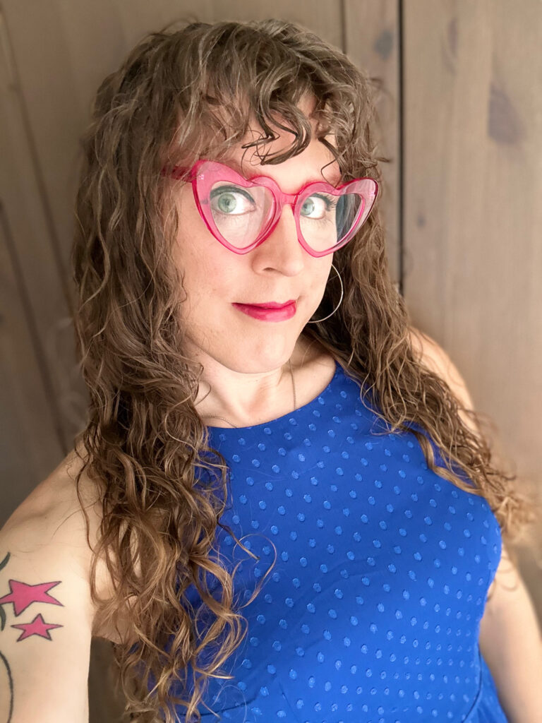 Me in a blue dress with pink heart-shaped glasses, dark eyeliner, and dark pink lipstick. I have long brown curly hair and curly bangs. And I look HAPPY. Because I am.