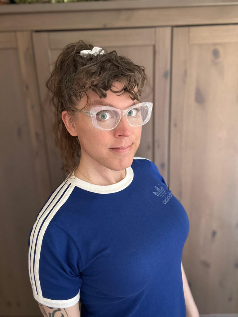 Me! with brown curly hair in a high ponytail and curly bangs, wearing a sporty short-sleeved blue ringer T, with a white collar and arm cuffs, and white stripes on the shoulders.