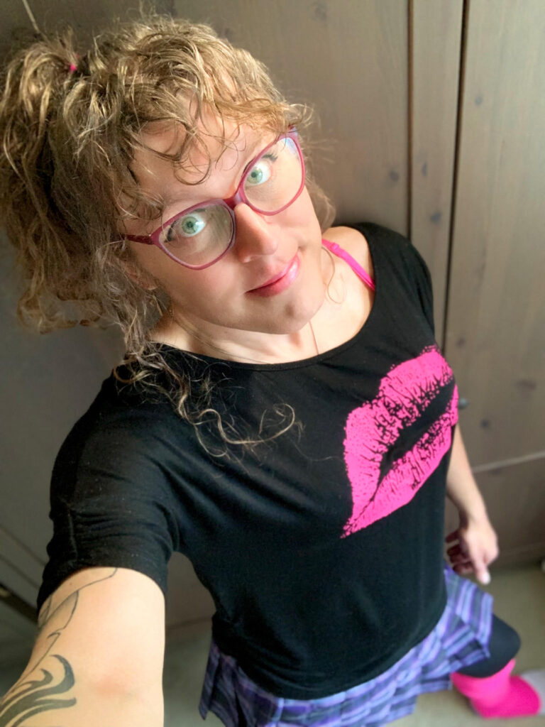 Me in a black cold shoulder top with a hot pink lipstick kiss on the front and one hot pink bra strap showing, a purple and pink pleated skirt, black tights, and hot pink legwarmers, with my long brown curly hair up in a pineapple