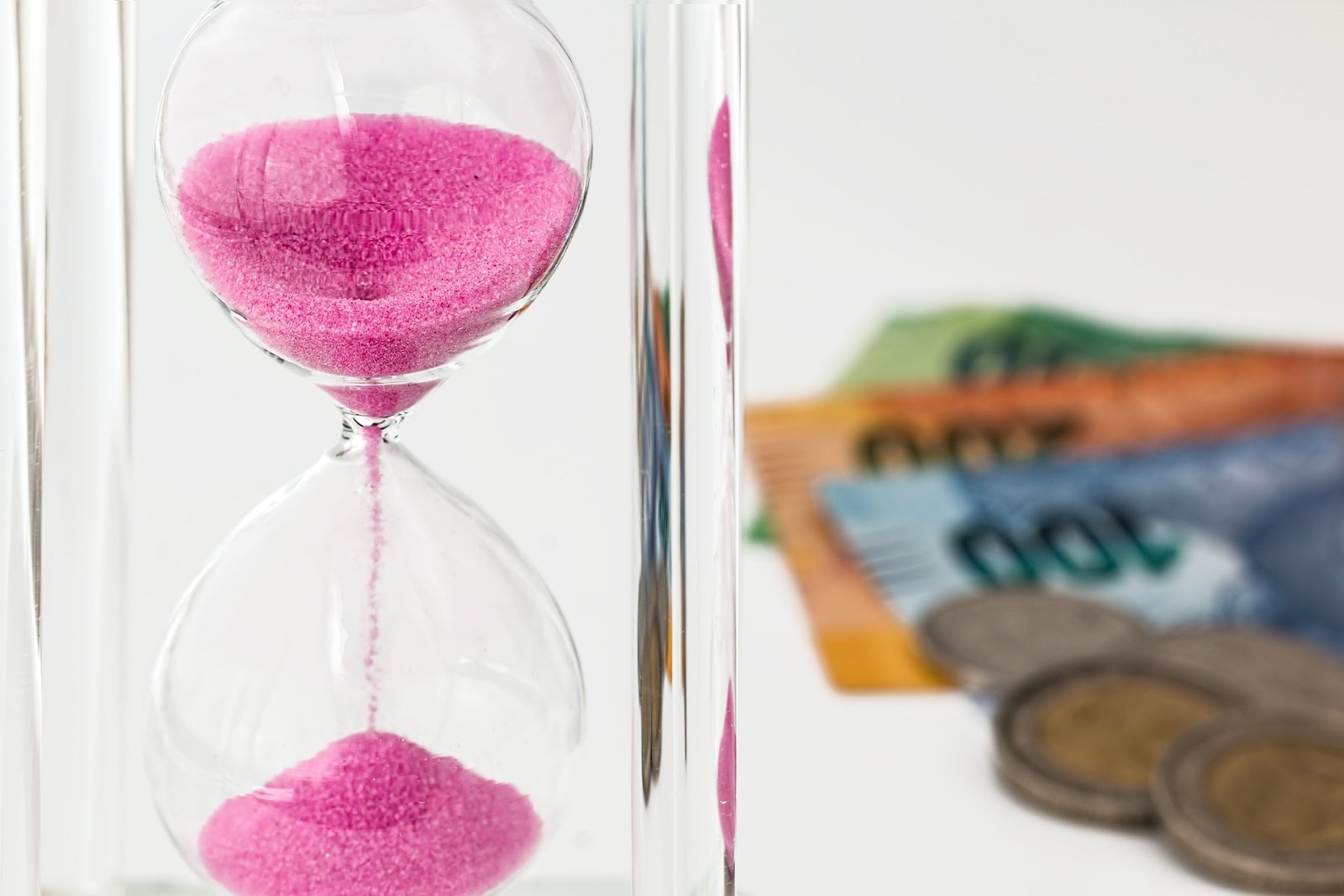 Fuchsia sand running through an hourglass. To its right is a pile of colorful money.