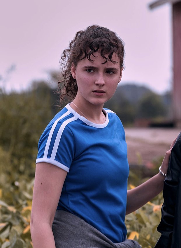 Fina Strazza as KJ, with brown curly hair in a high ponytail and curly bangs, wearing a sporty short-sleeved blue ringer T, with a white collar and arm cuffs, and white stripes on the shoulders.