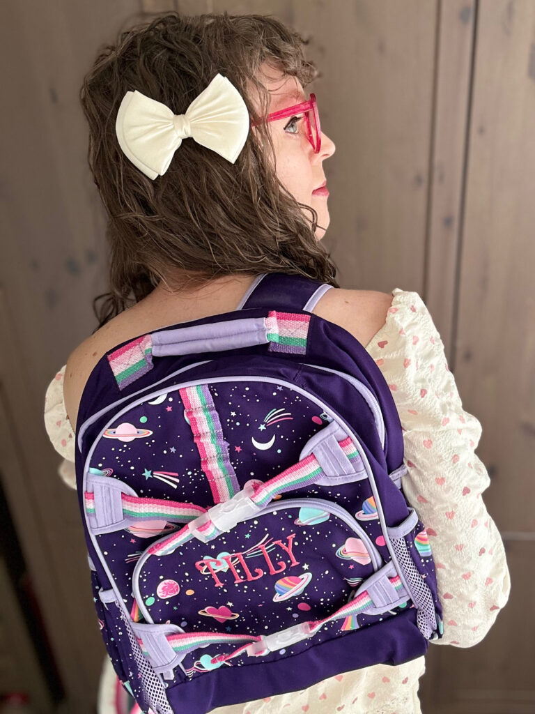 a purple and pink backpack with a girly-colored outer space print slung over one of my shoulders. Some of the planets and comets are replaced with hearts, and the straps are kind of pastel-colored rainbows.