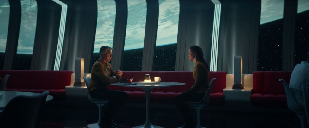 Pike and Batel talking while seated at a table in front of large windows, showing Earth outside the ship