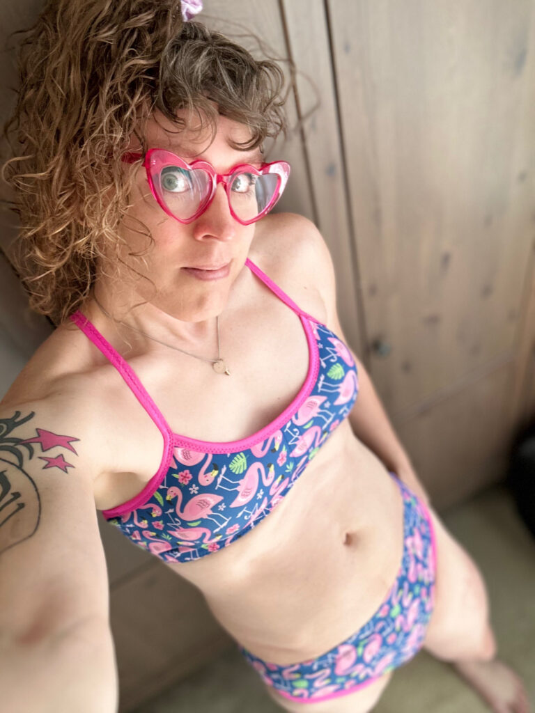 me in a blue bikini with hot pink trim and pink flamingos on it, my long brown curly hair is up in a pineapple and i have pink heart-shaped glasses