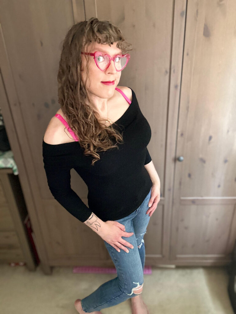 Me in an off the shoulder long sleeve black top with hot pink bra straps showing, hot pink lipstick and hot pink heart shaped glasses, and artfully ripped jeans, with long brown curly hair and curly bangs, and you can see the (tiny) curve of my hips!!