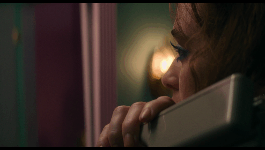 Side closeup of Lisa crimping her hair, a yellow light on the wall in the background, on a green-blue wall, right in front of her eyes as she looks into a mirror with a pink frame.