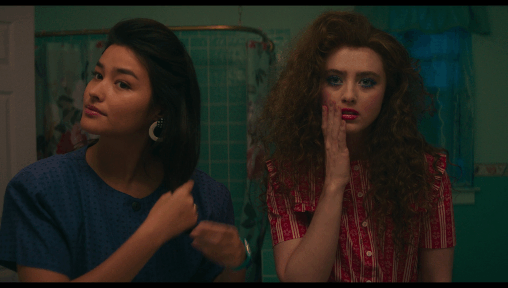 Taffy fixing her hair as she looks in a mirror. Lisa stands next to her, looking into the mirror, gently touching her face, looking miserable.