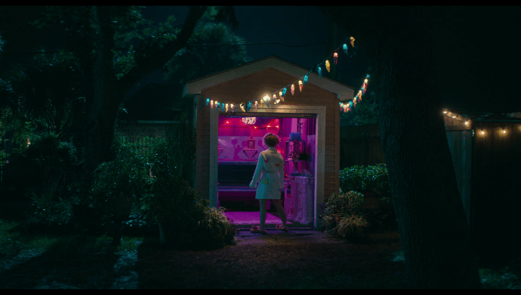 Lisa stands in a bathrobe in front of the shed with the tanning bed in it, the interior all pink and blue. Strings of yellow, red, and blue lights lead toward the shed.