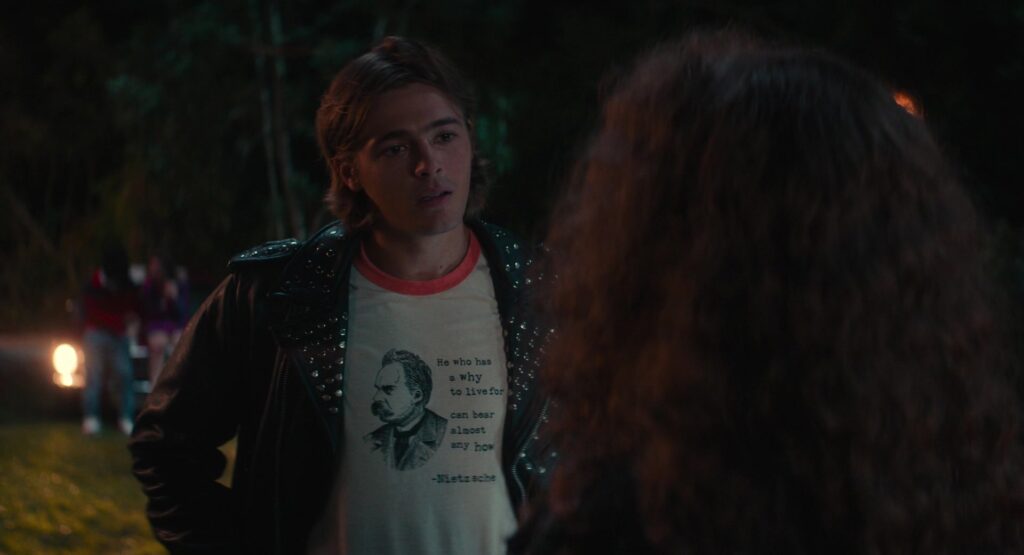 
Michael wears a white tee with an orange collar under a black studded leather jacket. The shirt has a picture of Nietzsche on it, and says “he who has a why to live for can bear almost any how”
