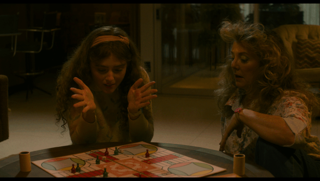 Lisa, in a white sweatshirt with pink flowers on it and a pink headband, sits on the floor playing a board game with her mom. The spaces on the board are pink and blue, green and yellow, red, and the pieces are red and blue.