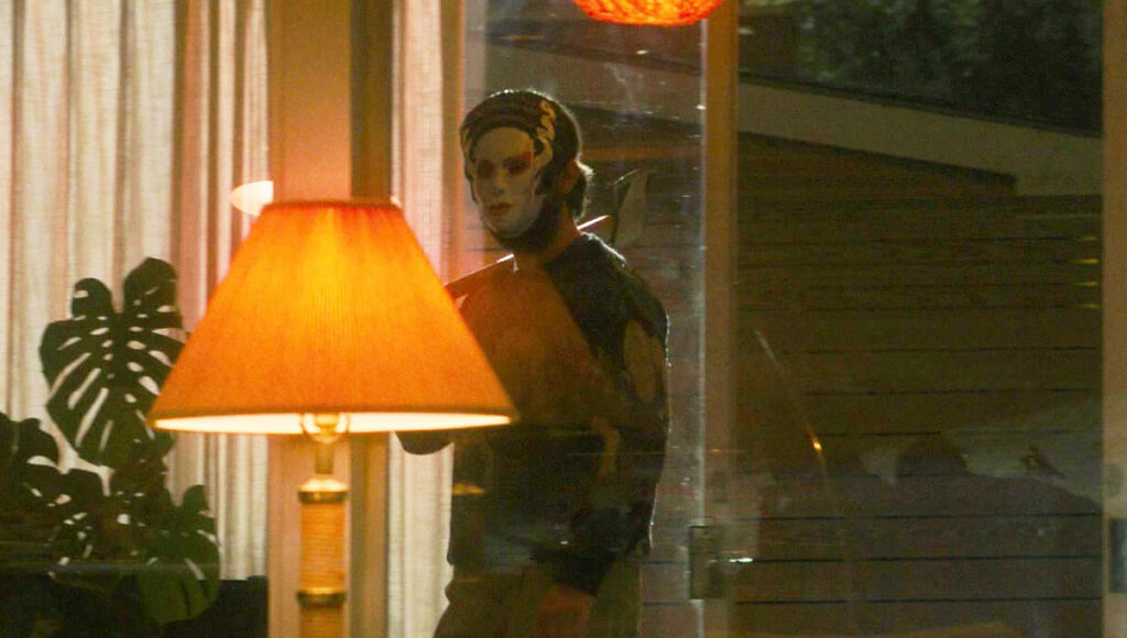 A man in a bride of Frankenstein mask enters a house behind a red lamp, he’s mostly in darkness, with the image lightened so you can see the man in shadow better.