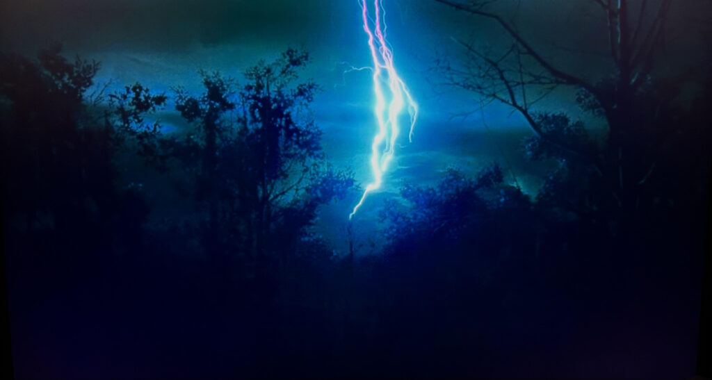 Pink, white, and blue lightning strikes in the forest, in front of a green-blue sky