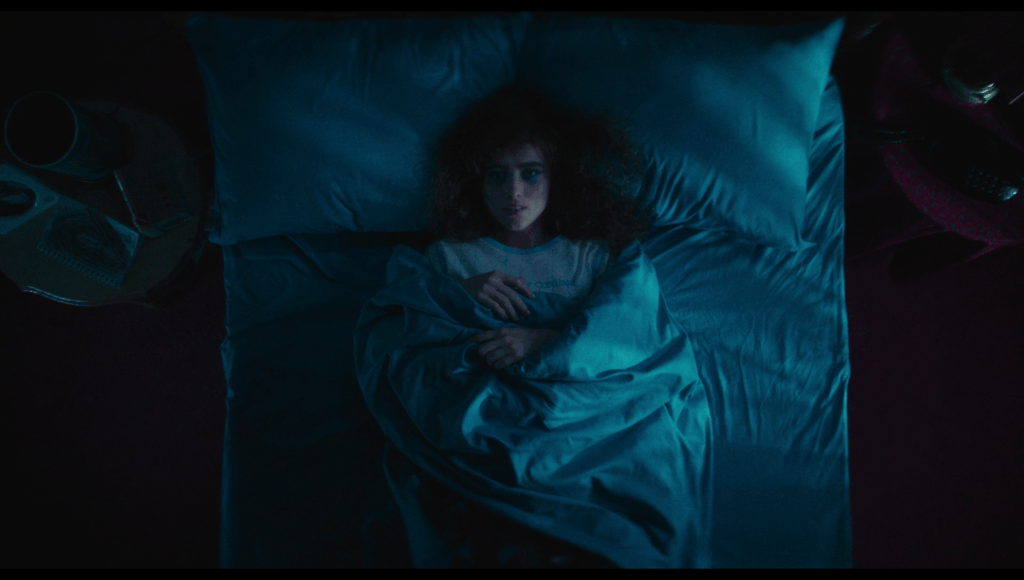 Lisa, wrapped up in her blue blankets in her blue bed, shadows from vertical blinds at the window casting shadows across her bed like prison bars.