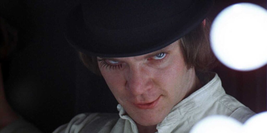 A young Macolm McDowell in A Clockwork Orange, with one false eyelash on the bottom of his right eye, head down but eyes up.