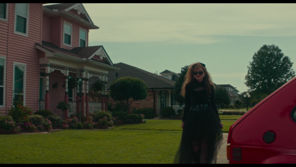 Lisa in a floor length black dress with long sleeved and dark sunglasses walks toward taffy’s car.