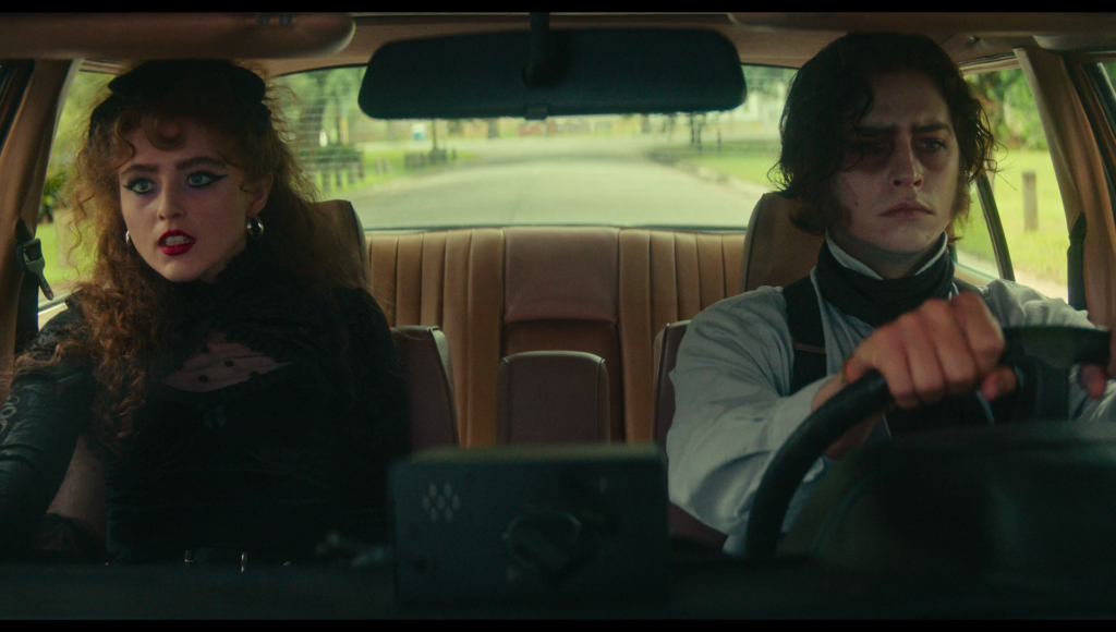 Lisa and Frank sitting side by side, Frank driving, in a yellow car. Both wear semi-formal Victorian-style clothing.