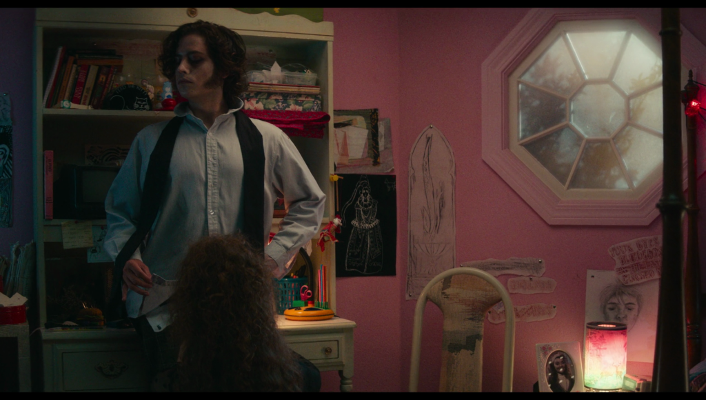 Lisa kneels on the floor in front of a standing Frank, next to the large penis drawing on the wall.