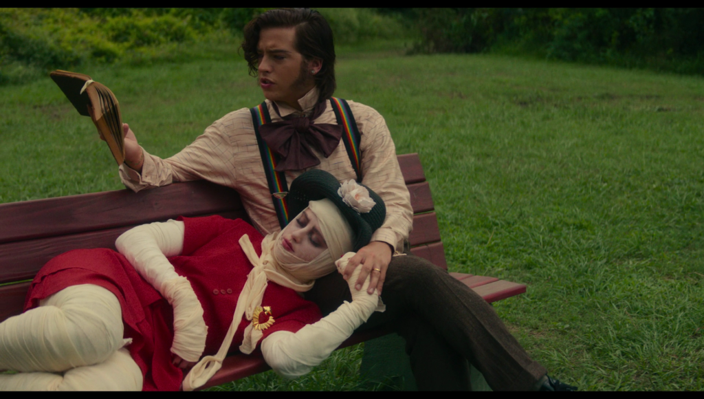 Lisa, in a red dress over an entirely bandaged body, with a pale white face and a red heart brooch, lays on the red bench with her head in Frank’s lap, as he reads to her from a book. Frank has rainbow suspenders on.