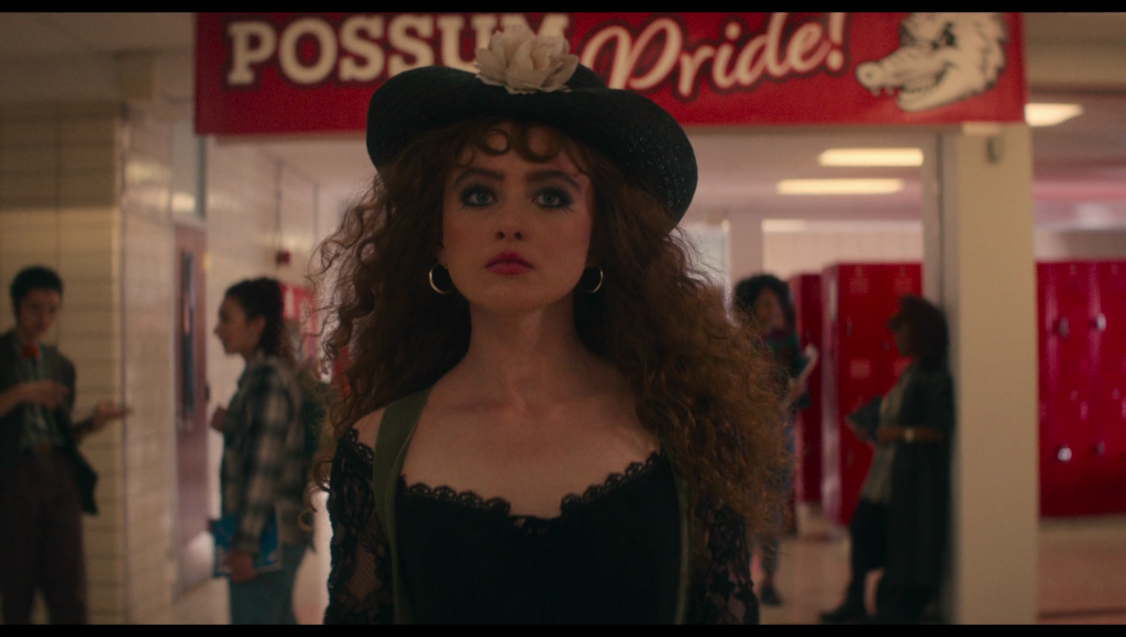 Lisa in a black dress and black hat, surrounded by red lockers and a red banner that reads “possum pride”