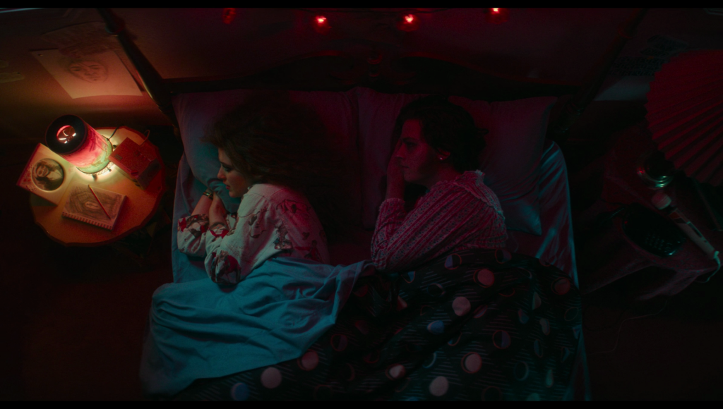 Lisa and Frank lying next to each other in bed, in the same positions, both facing a pink and blue lamp on the bedside table.