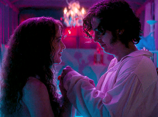 animated gif from Lisa Frankenstein of Lisa and the creature/Frank kissing each other's hands under pink and blue lighting