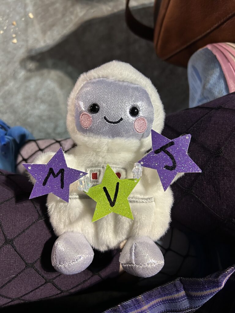 a small stuffed astronaut holding a string of stars that say M V J