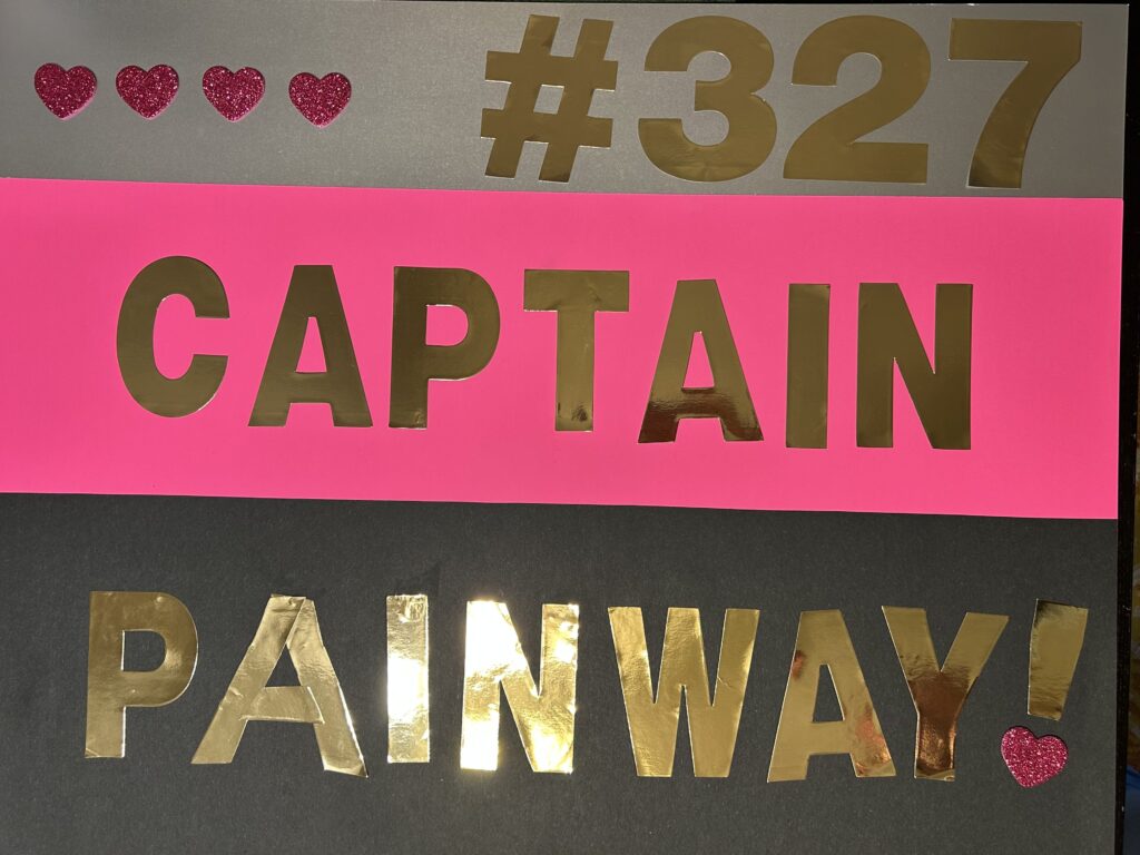 three horizontal bars, grey on top, pink in the middle, and black on bottom, resembling the top of a Star Trek voyager uniform. Where the rank pips would be on the gray bar are four sparkly pink hearts, and on the right side in gold is #327. on the pink middle bar is “Captain” in gold letters, and on the bottom black bar is “Painway!” In gold letters, and the dot under the exclamation point is another sparkly pink heart