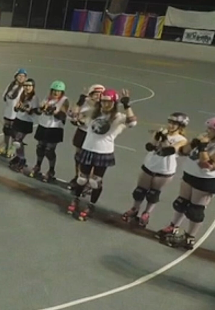 Me in a line of roller derby players, on a rink, waving. On the fence to the rink in the background are a line of pride flags.
