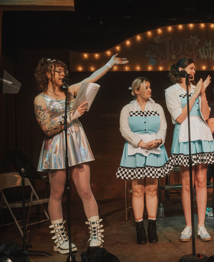 Me in a holographic shimmery dress with holographic shimmery bows in my hair, white fishnet gloves, sparkly white fishnets, and buckled white platform boots with reflective stars on them, holding a script and standing at a microphone, gesturing to some of the cast next to me as I read out the credits