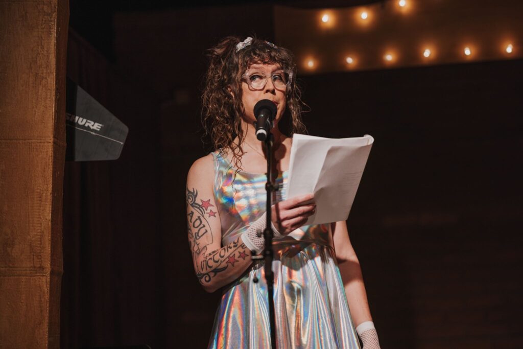 Me in a holographic dress, holding a script, on stage speaking into a microphone