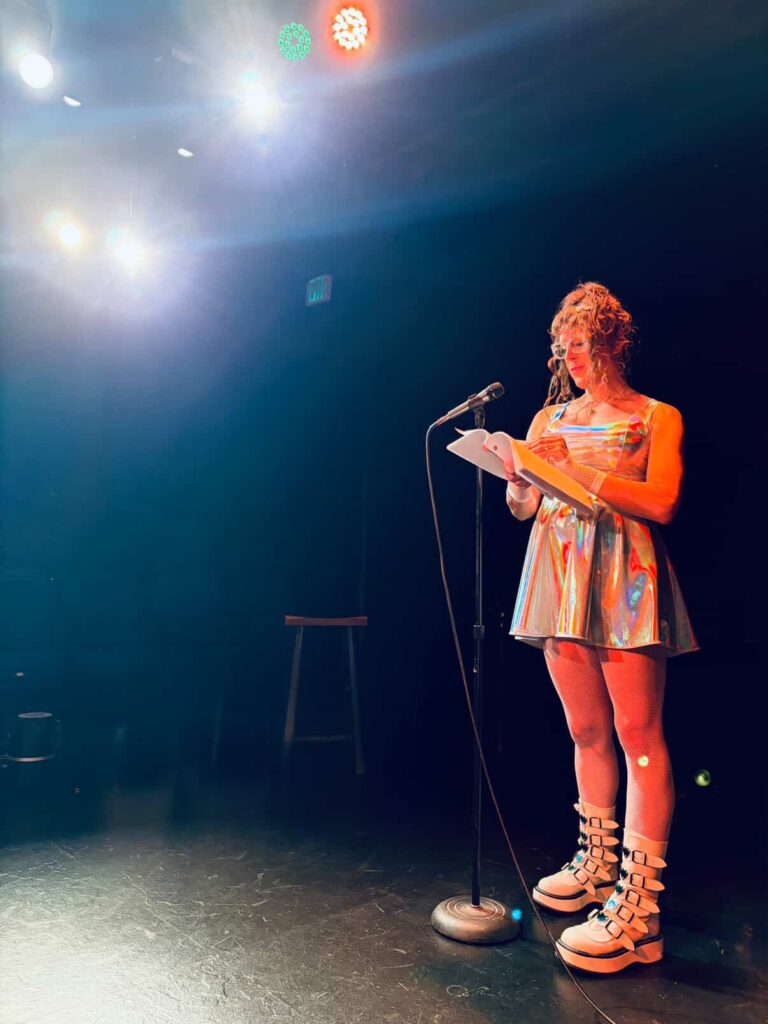 Me in a holographic shimmery dress with holographic shimmery bows in my hair, white fishnet gloves, sparkly white fishnets, and buckled white platform boots with reflective stars on them, holding a script and standing at a microphone