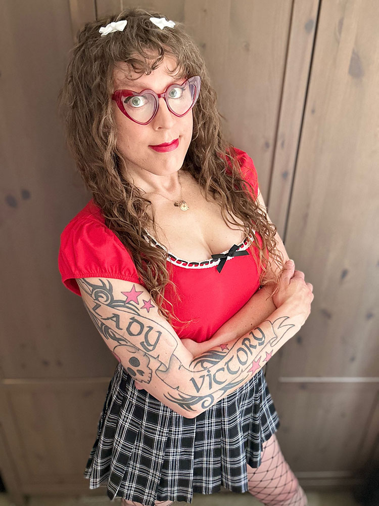 me in a red top with white lace trim and a black bow, with my arms folded under my boobs, with red heart glasses and white bows in my long brown curly hair, with a black and white plaid pleated skirt, and black fishnets